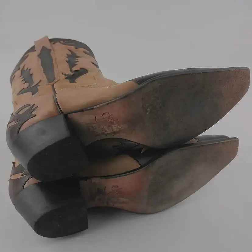 A pair of cowboy boots shown from the back, featuring tan leather shafts with black western-style embroidery and black leather heels, set against a plain light gray background.