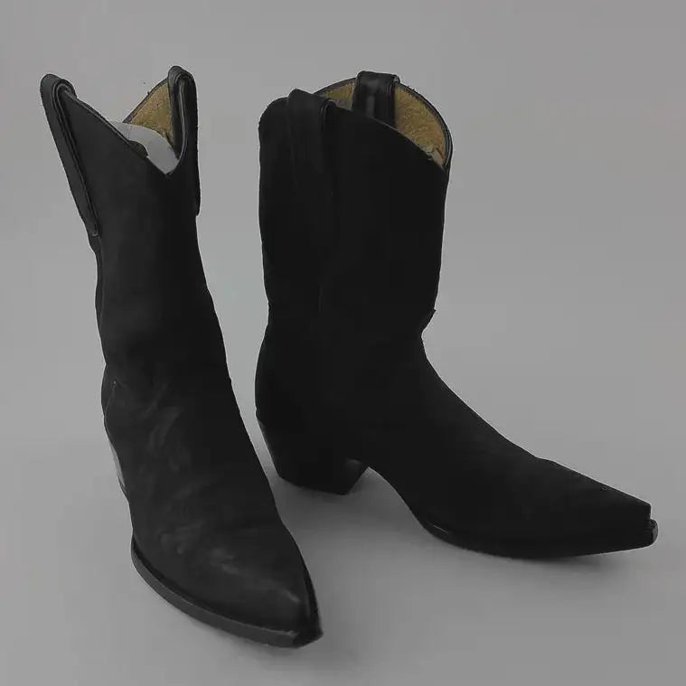 A pair of black leather cowboy boots with pointed toes and low block heels, photographed on a neutral gray background.