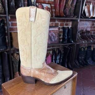 A light beige western cowboy boot with textured embossed detailing on the shaft and a tan textured leather vamp, displayed on a wooden cabinet in a boot store with brick walls and other boots in the background.