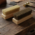 Two wooden cleaning brushes with light nylon and dark horsehair bristles resting on a rustic wooden workbench, with leatherworking tools in the background.