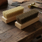 Two wooden cleaning brushes with light nylon and dark horsehair bristles resting on a rustic wooden workbench, with leatherworking tools in the background.
