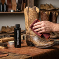 A hand wipes a distressed brown leather cowboy boot with a red cloth next to a bottle of Boot Collector's boot care conditioner product on a wooden workbench.