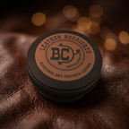 A round black tin of Leather Nourisher by Boot Collector, featuring a bronze circular label, resting on a textured brown leather surface with warm bokeh lights in the background.