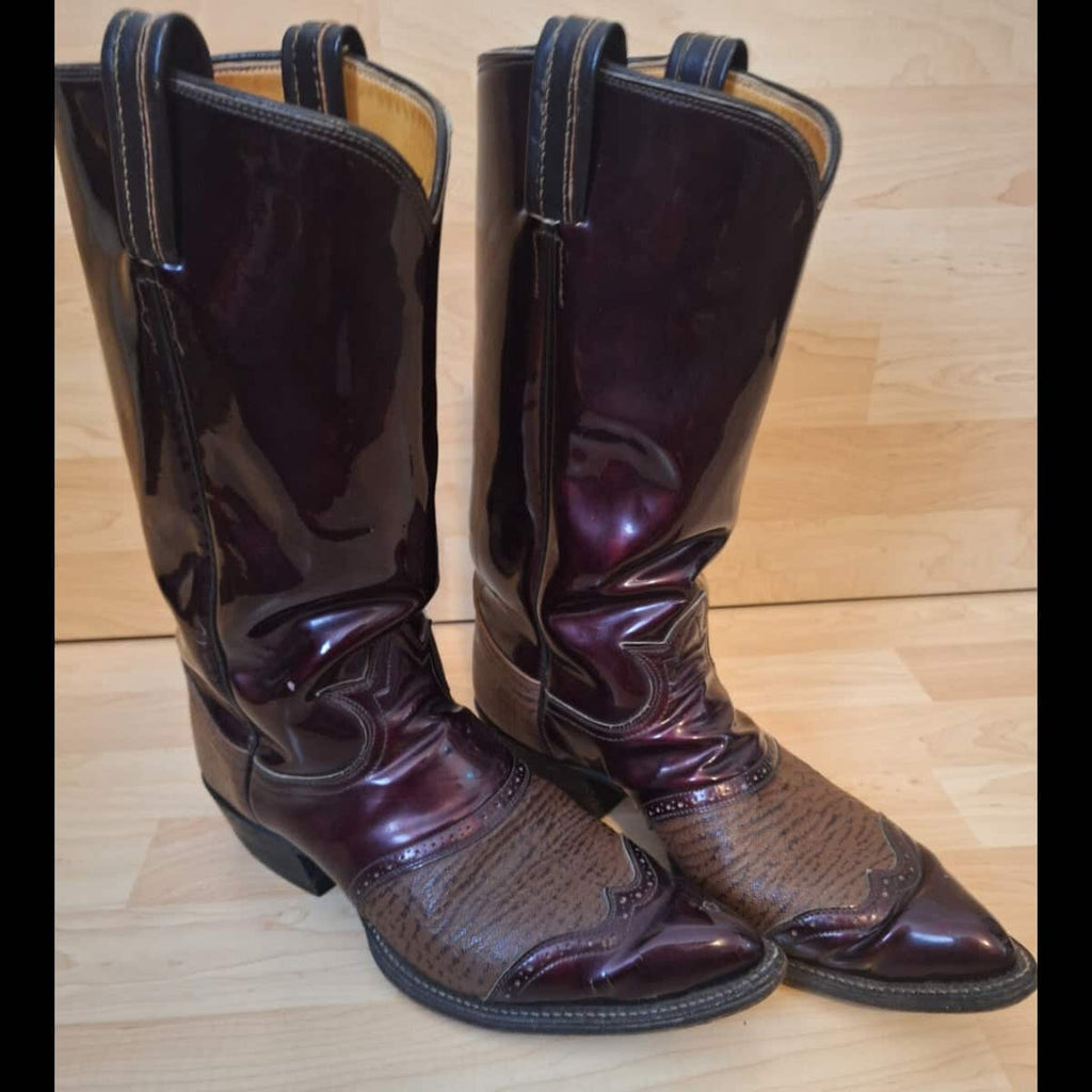 A pair of women's western cowboy boots in a glossy dark burgundy patent leather with a textured brown brogue toe cap, displayed on a light wood floor.