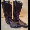 A pair of women's western cowboy boots in a glossy dark burgundy patent leather with a textured brown brogue toe cap, displayed on a light wood floor.