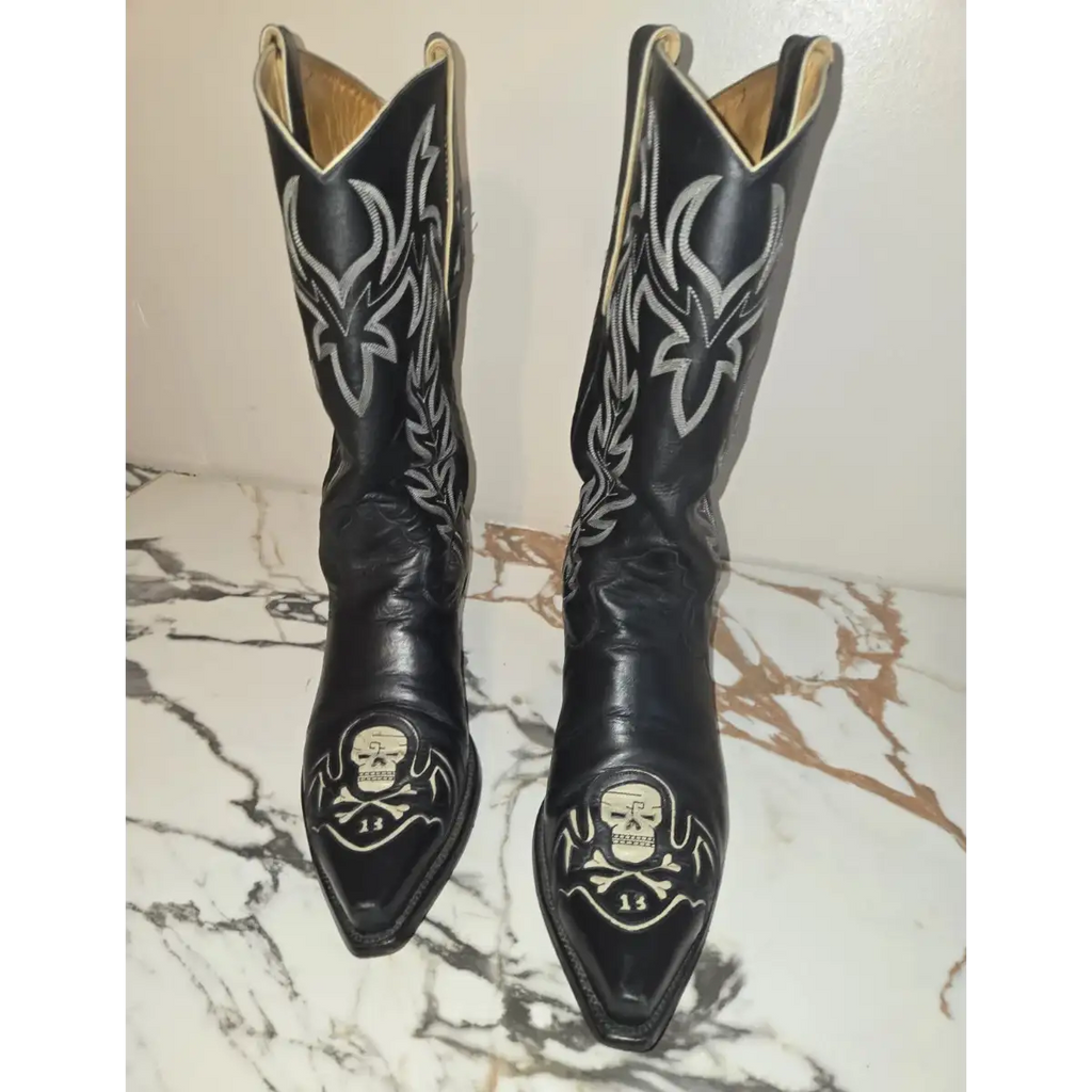 A pair of black leather cowboy boots with silver western embroidery and skull designs on the toes, displayed on a white and brown marble surface against a plain wall.