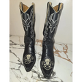A pair of black leather cowboy boots with silver western embroidery and skull designs on the toes, displayed on a white and brown marble surface against a plain wall.