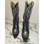 A pair of black leather cowboy boots with silver western embroidery and skull designs on the toes, displayed on a white and brown marble surface against a plain wall.