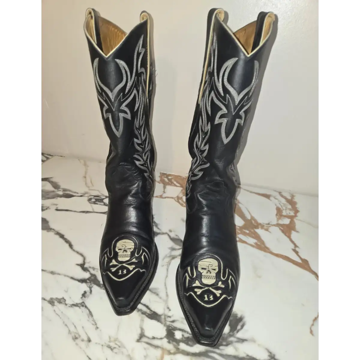 A pair of black leather cowboy boots with silver western embroidery and skull designs on the toes, displayed on a white and brown marble surface against a plain wall.