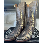 A pair of distressed brown leather cowboy boots with metallic gold and black textured fringe detailing, displayed on a black shoebox in a living room setting.