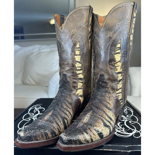 A pair of distressed brown leather cowboy boots with metallic gold and black textured fringe detailing, displayed on a black shoebox in a living room setting.