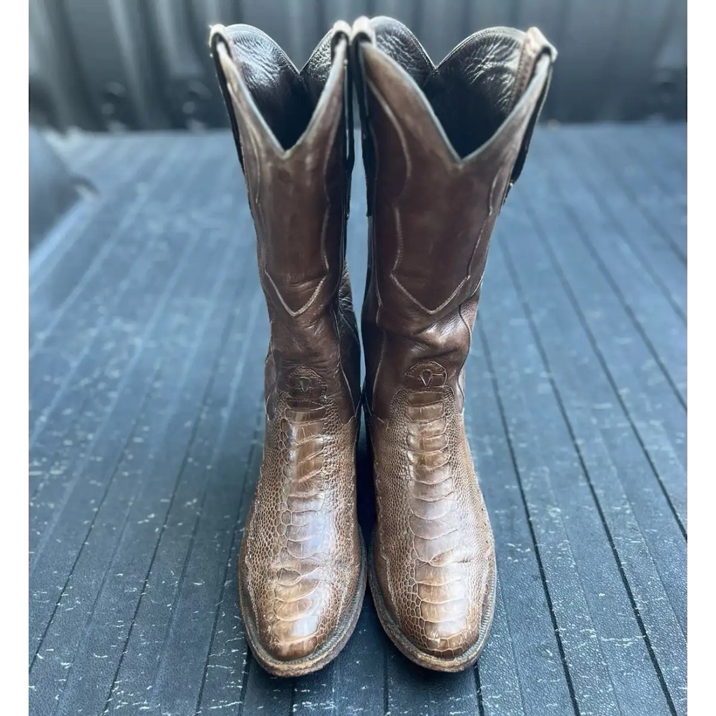 A pair of brown leather cowboy boots with a textured alligator or crocodile skin pattern, standing upright on a dark gray wooden surface.