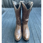 A pair of brown leather cowboy boots with a textured alligator or crocodile skin pattern, standing upright on a dark gray wooden surface.
