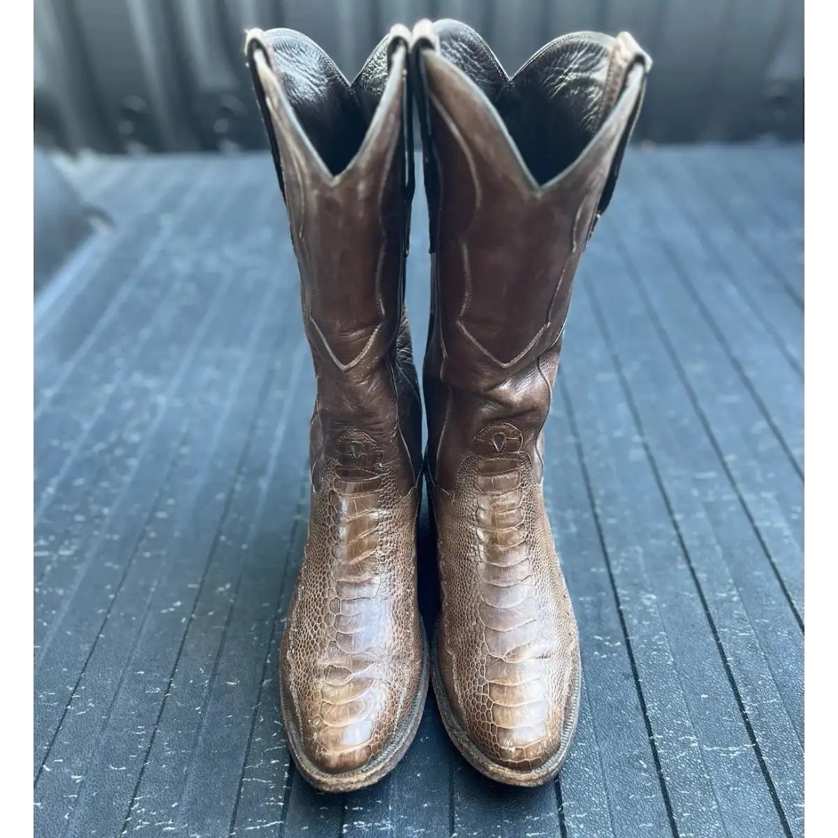A pair of brown leather cowboy boots with a textured alligator or crocodile skin pattern, standing upright on a dark gray wooden surface.