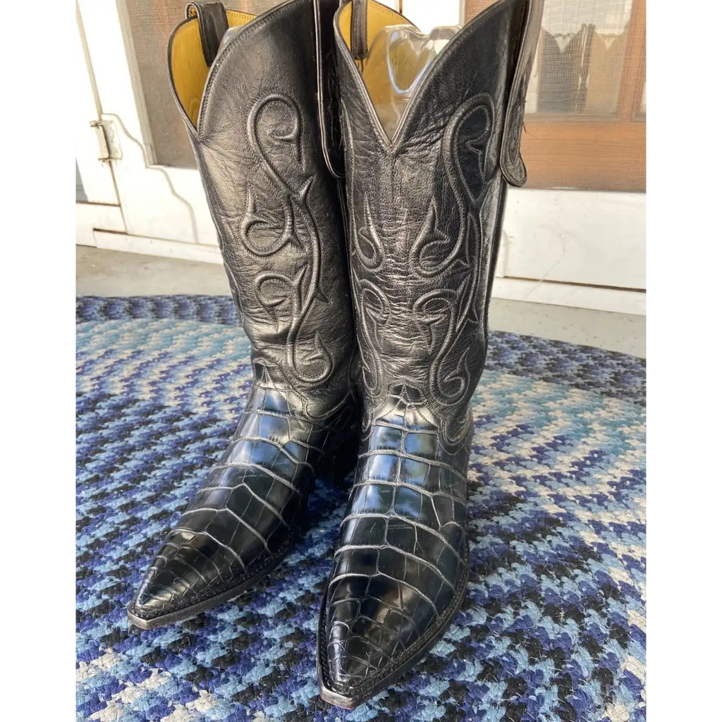 A pair of black alligator-textured cowboy boots with carved western shaft detailing, standing on a blue and white patterned rug in front of a glass door.