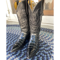 A pair of black alligator-textured cowboy boots with carved western shaft detailing, standing on a blue and white patterned rug in front of a glass door.