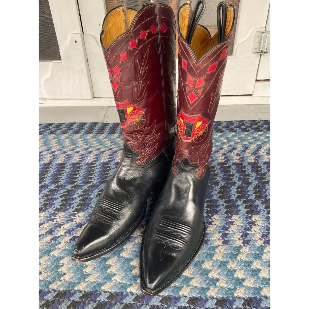 A pair of cowboy boots with black leather lower shafts and red leather upper shafts decorated with geometric and floral embroidery, standing on a blue and white patterned rug.