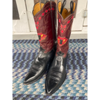 A pair of cowboy boots with black leather lower shafts and red leather upper shafts decorated with geometric and floral embroidery, standing on a blue and white patterned rug.