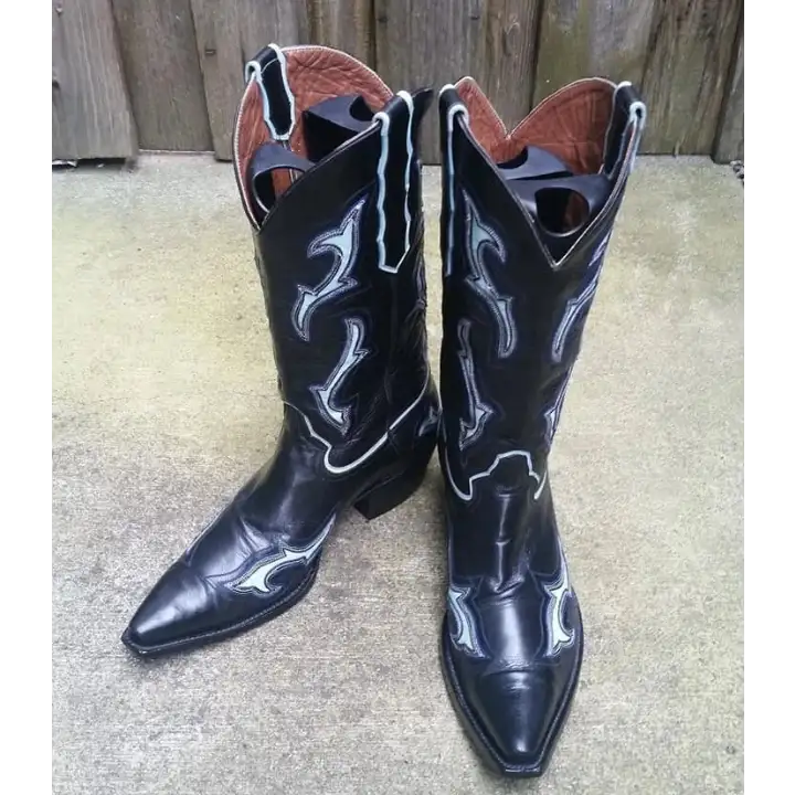 A pair of black leather cowboy boots with white western-style stitching, standing on a concrete surface in front of a wooden fence.