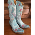 A pair of light blue western cowboy boots with intricate multicolor embroidery featuring floral and folk-art motifs, displayed on a wooden floor against a wooden wall.