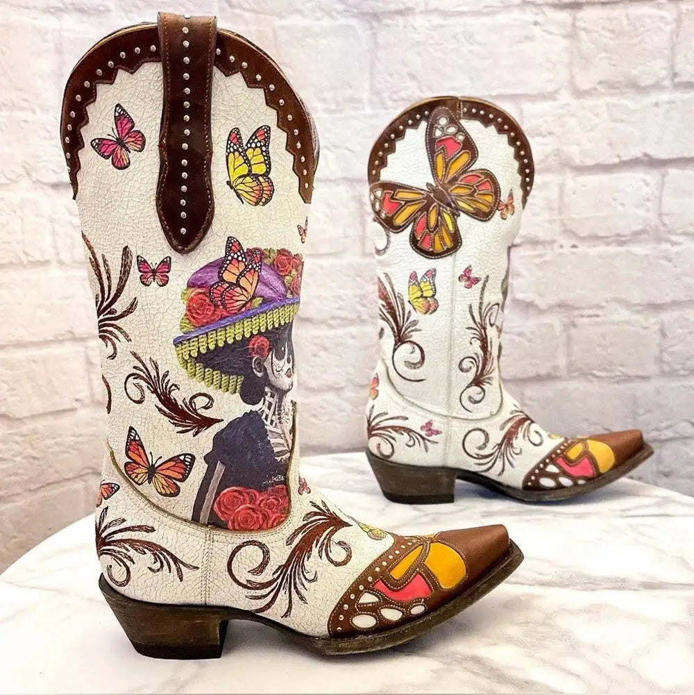 A pair of hand-painted white leather cowboy boots with brown leather trim, decorated with colorful butterflies, floral motifs, and a Day of the Dead skeleton design, displayed on a white marble surface against a white brick wall.