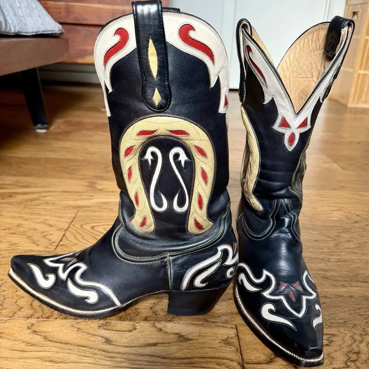 A pair of black leather cowboy boots with white, red, and beige embroidered western designs, standing on a wooden floor.