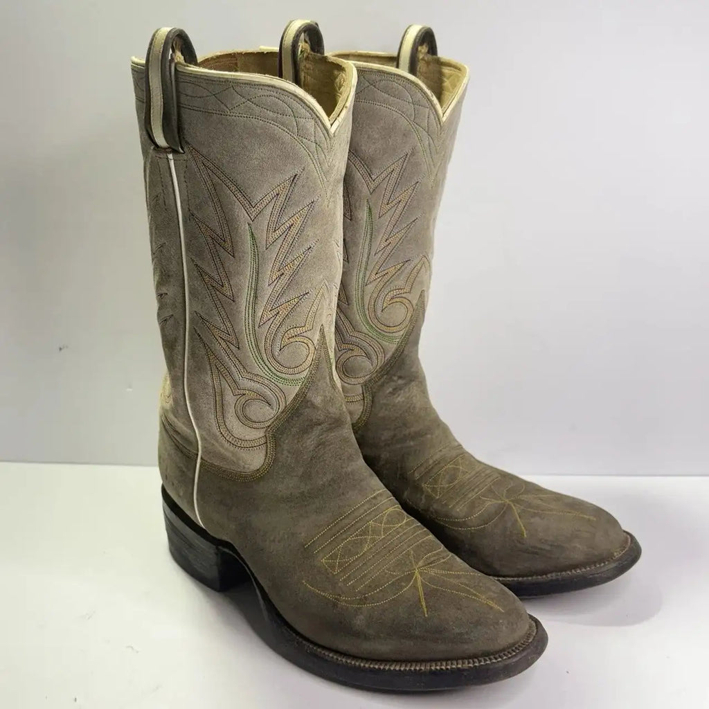 A pair of western cowboy boots in a distressed taupe-brown leather, featuring decorative embroidered stitching in tan and light green, set against a plain light gray background.