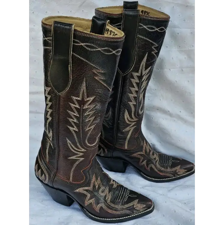 A pair of dark brown western cowboy boots with tan and beige embroidered floral and geometric patterns, displayed on a light gray fabric background.