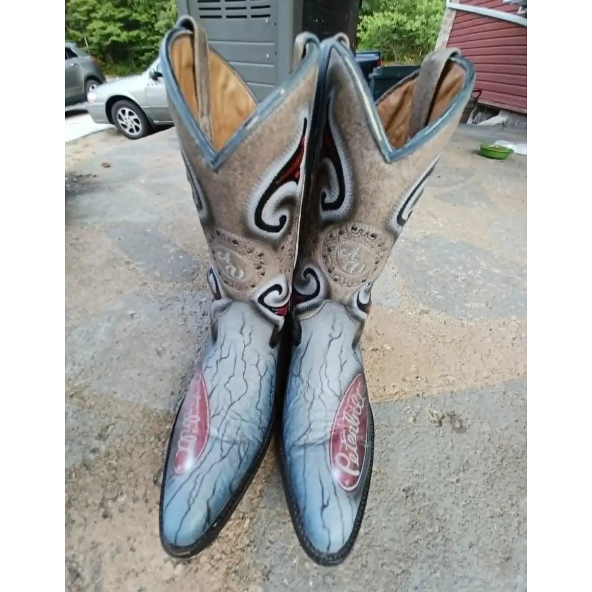 A pair of western cowboy boots with a cracked blue and gray leather finish, featuring black and white scrollwork embroidery and red 'Corning' glass on the toes, standing on a concrete driveway.