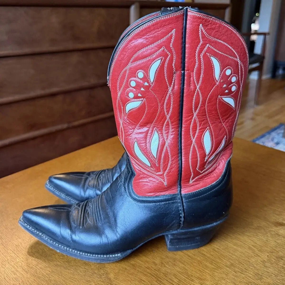 A pair of cowboy boots with red western shafts featuring white embroidered floral patterns and black leather feet, resting on a wooden table in an indoor setting.
