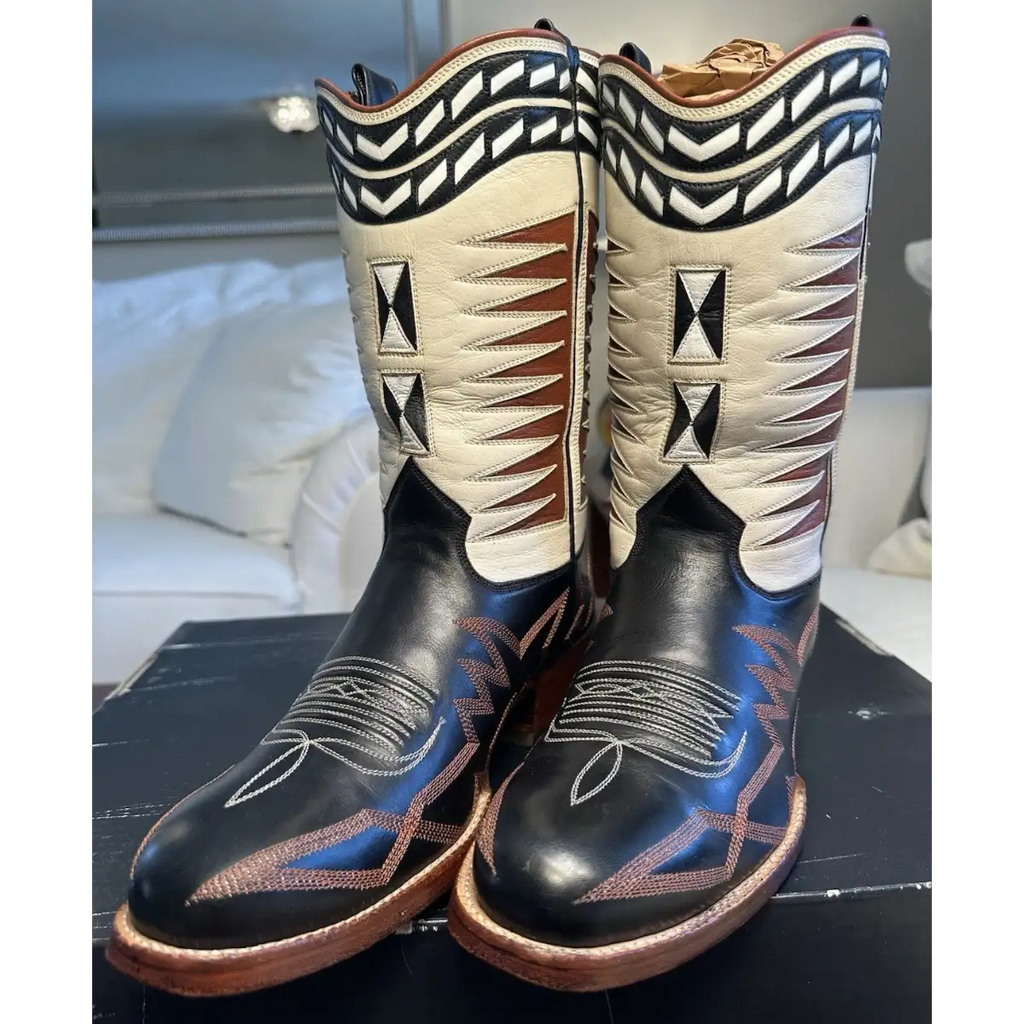 A pair of western cowboy boots with black leather lower sections, cream upper shafts, and intricate brown, black, and blue decorative stitching, displayed on a black surface with a blurred bedroom background.