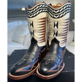 A pair of western cowboy boots with black leather lower sections, cream upper shafts, and intricate brown, black, and blue decorative stitching, displayed on a black surface with a blurred bedroom background.