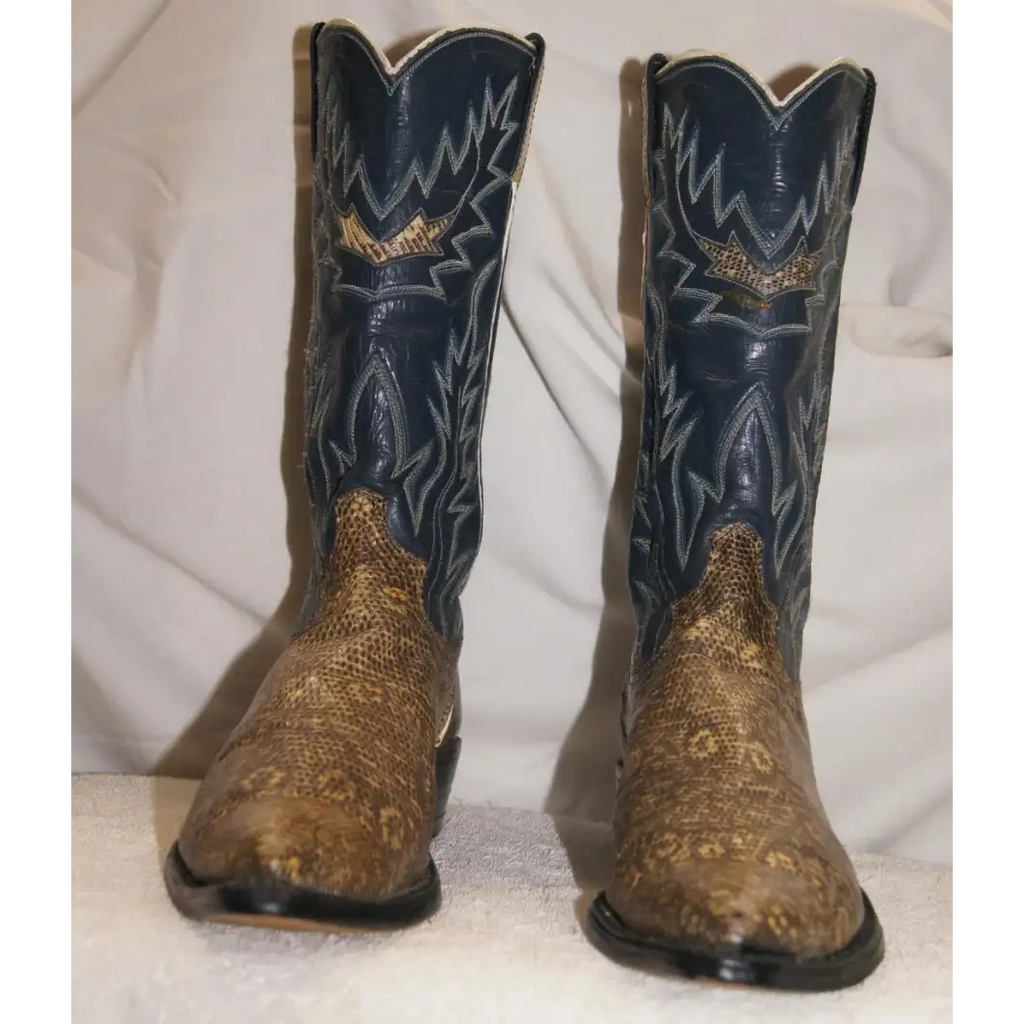 A pair of cowboy boots with dark navy blue shafts featuring gold western embroidery and a brown snakeskin-patterned foot, set against a draped white fabric background.