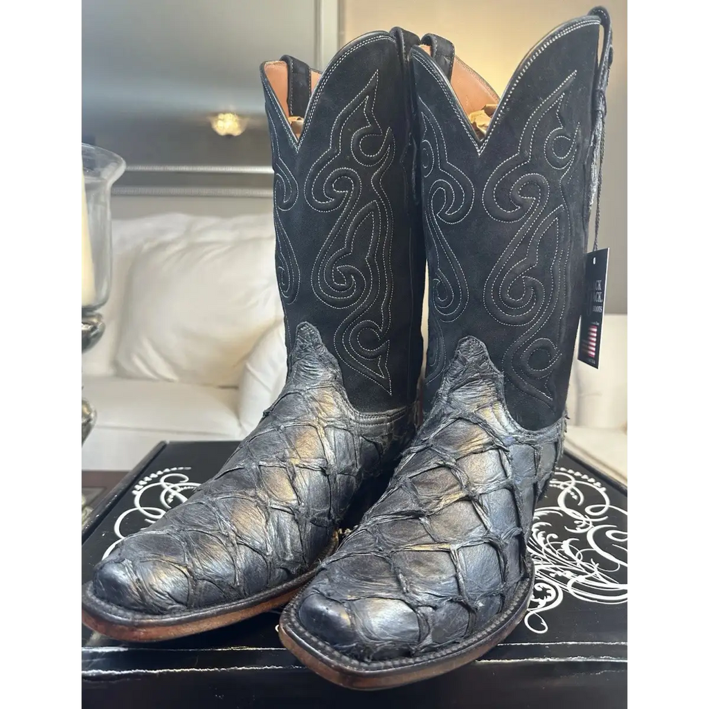 A pair of black western cowboy boots with textured alligator or crocodile skin on the lower shafts and decorative contrast stitching, displayed on a black box in a home setting.