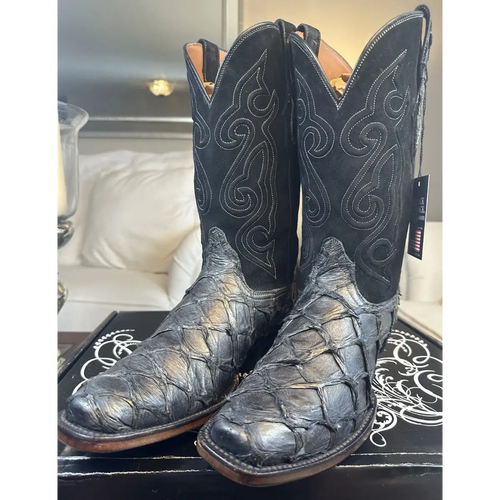 A pair of black western cowboy boots with textured alligator or crocodile skin on the lower shafts and decorative contrast stitching, displayed on a black box in a home setting.