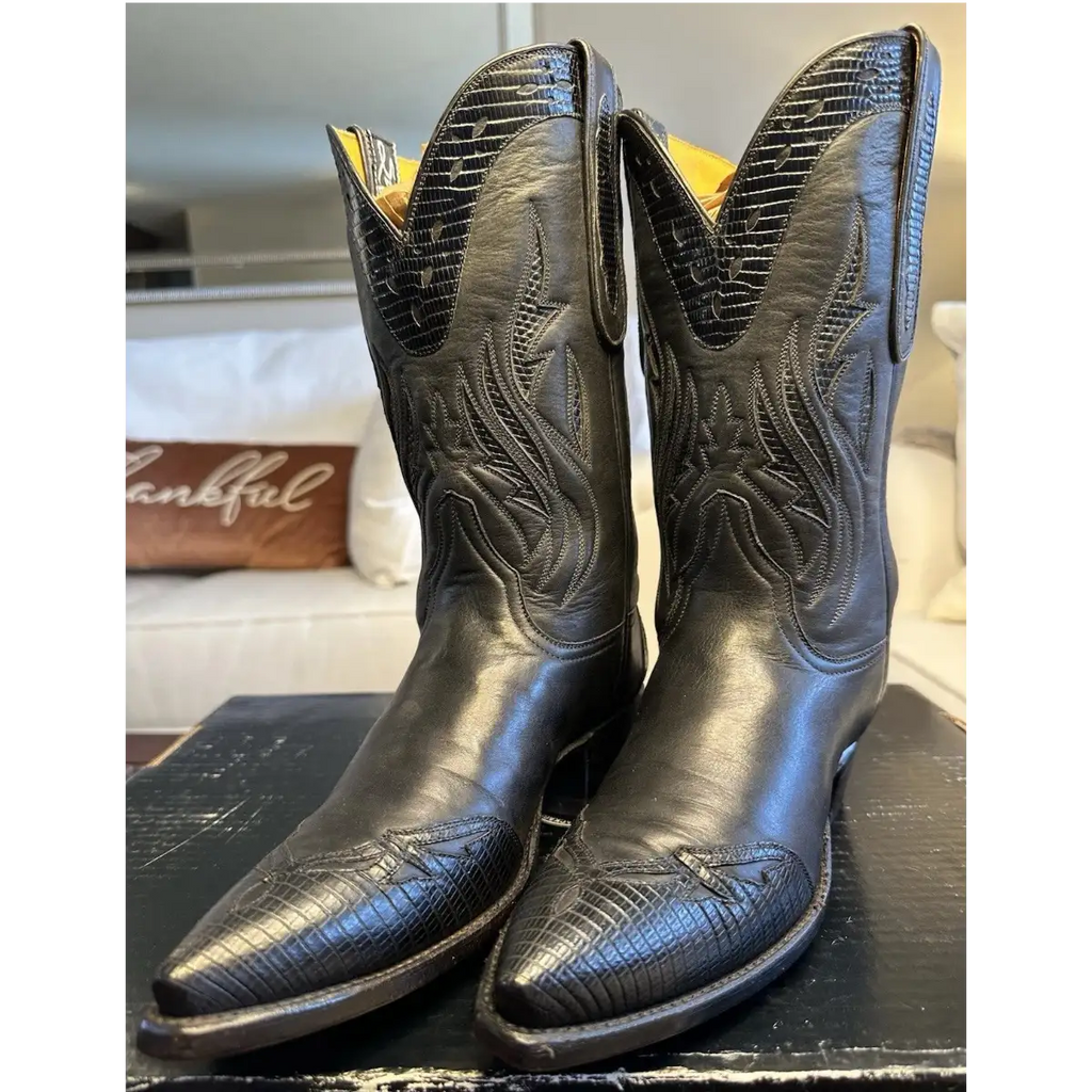 A pair of black western cowboy boots with embossed alligator-style texture and floral stitching, displayed on a dark wooden surface in a home setting.