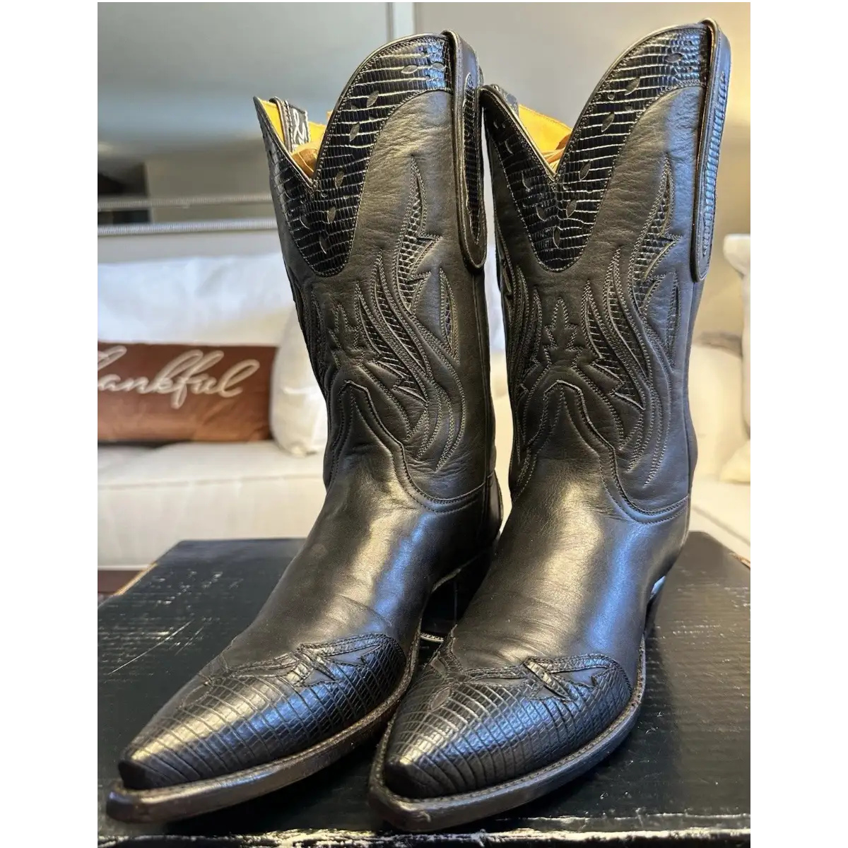 A pair of black western cowboy boots with embossed alligator-style texture and floral stitching, displayed on a dark wooden surface in a home setting.